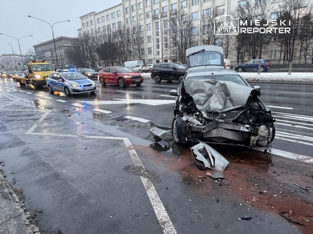 Zniszczony samochód osobowy na pasie ruchu, obok policyjny radiowóz oraz laweta, w tle ruch uliczny i budynki.