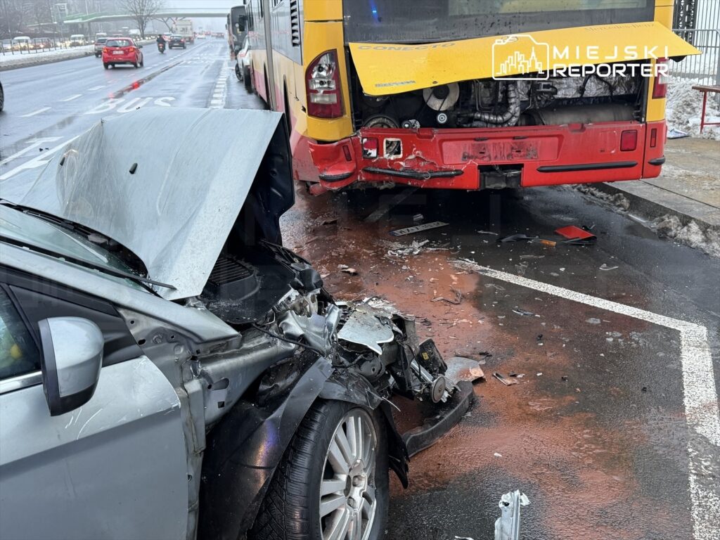 Zniszczony samochód osobowy uderzający w tył autobusu, na jezdni pokrytej błotem i resztkami oleju.