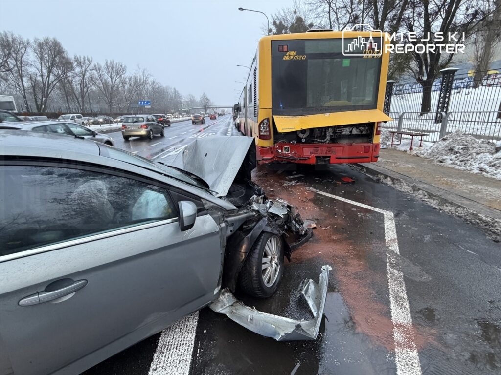Zderzenie samochodu osobowego z autobusem na ruchliwej ulicy w zimowej scenerii, z uszkodzeniami pojazdów i śladami płynów.