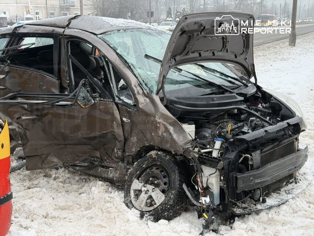 Uszkodzony samochód osobowy z otwartą maską, stojący na zaśnieżonej drodze, obok czerwono-żółta taśma ostrzegawcza.