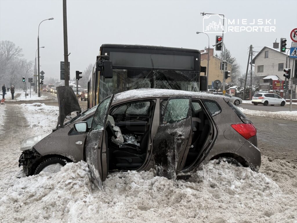 Samochód utknął w śniegu na skrzyżowaniu, obok zatrzymany autobus oraz inne pojazdy w zimowej scenerii.