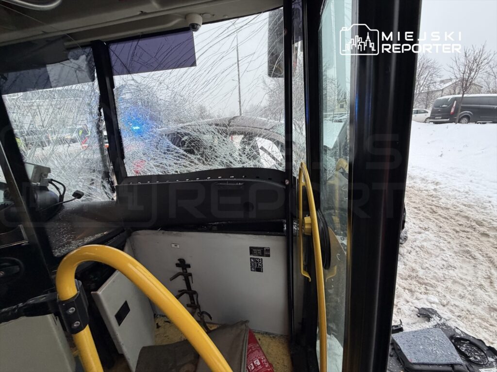 Zniszczona szyba w autobusie po kolizji, widok z wnętrza pojazdu, w tle policyjne światła i zaśnieżona droga.