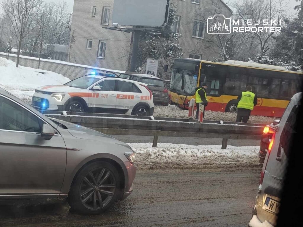 Policyjny radiowóz z niebieskimi światłami stoi obok autobusu miejskiego, a pracownicy w żółtych kamizelkach kierują ruchem w zaśnieżonej okolicy.