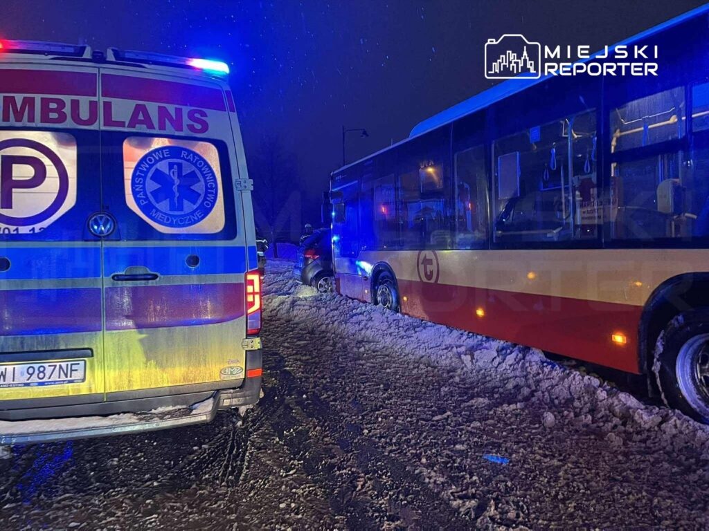 Ambulans z włączonymi sygnałami świetlnymi stoi na zaśnieżonej drodze obok unieruchomionego autobusu.