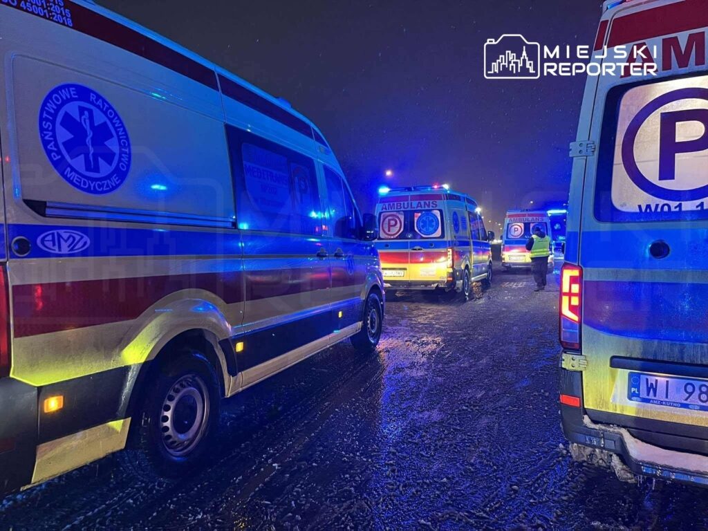 Sześć ambulansów z włączonymi sygnałami świetlnymi stoi na ośnieżonej drodze w nocy.