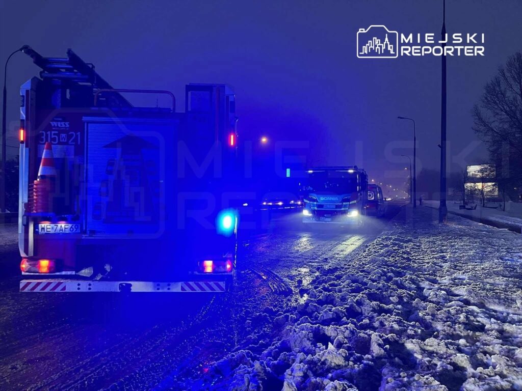 W nocy dwa pojazdy straży pożarnej z włączonymi sygnałami świetlnymi stoją na zaśnieżonej drodze.