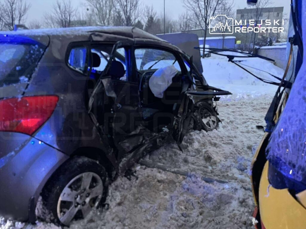 Uszkodzony samochód osobowy leży w śniegu po kolizji, obok widać pojazd ratunkowy z włączonymi sygnalizatorami.