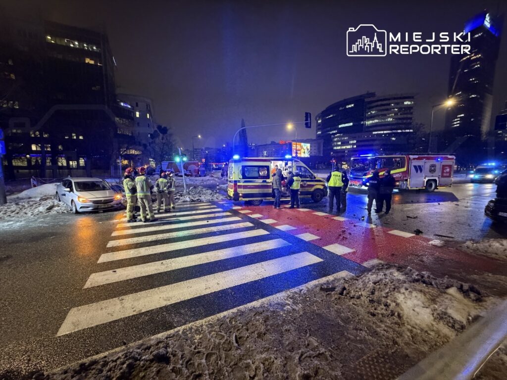 Grupa strażaków i policjantów pracuje na miejscu wypadku drogowego przy oświetlonym skrzyżowaniu w nocy.