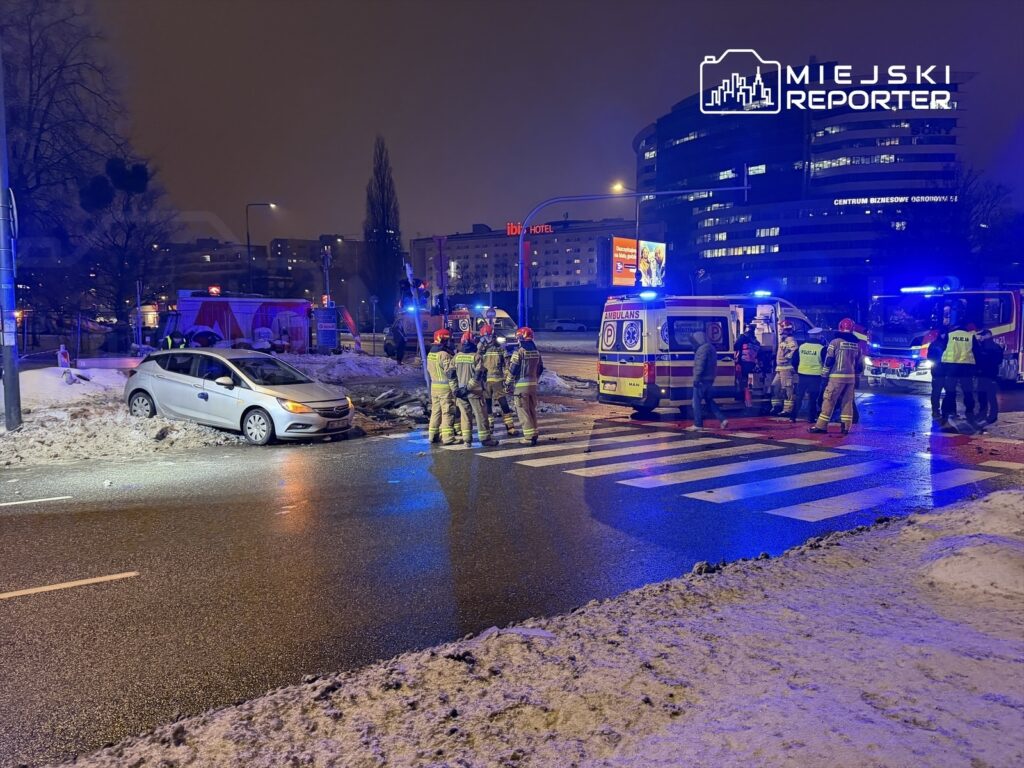 Biały samochód osobowy stoi na poboczu, a obok grupka strażaków i ratowników w akcji, w tle widać budynki i pojazdy służb.