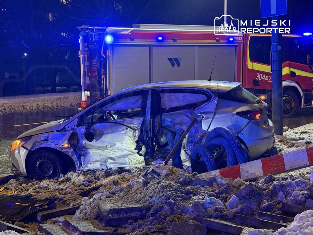 Rozbity samochód osobowy z otwartymi drzwiami stoi na zasypanej śniegiem jezdni, obok widać woz strażacki z włączonymi sygnałami.