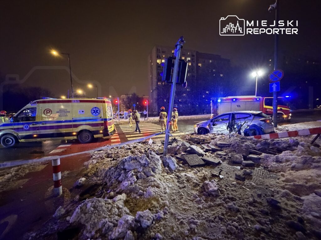 Na skrzyżowaniu nocą, ambulans i strażacy udzielają pomocy poszkodowanemu w wypadku samochodowym.