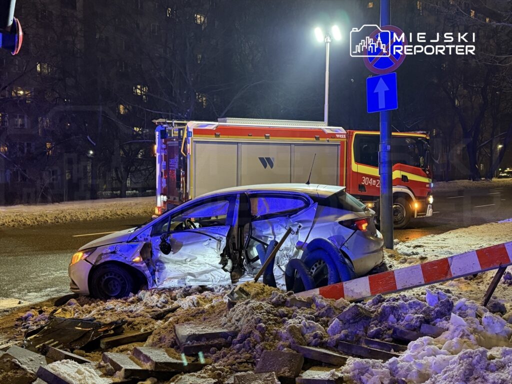 Rozbity samochód osobowy z otwartymi drzwiami znajduje się na zasypanej śniegiem jezdni obok wozu strażackiego.