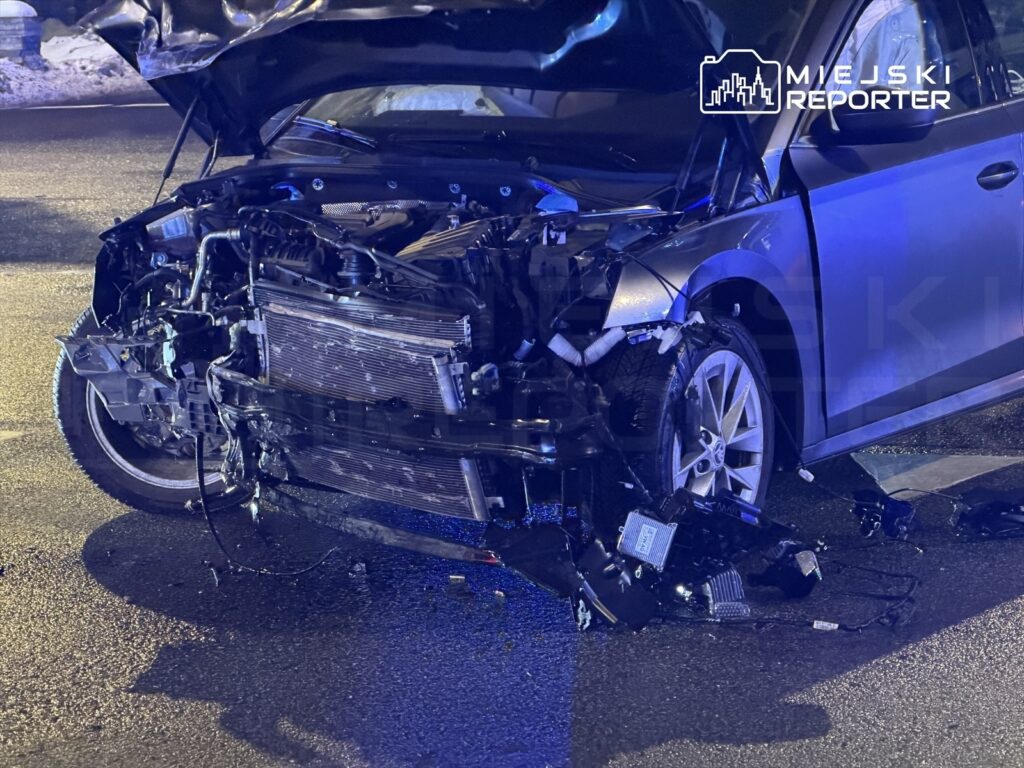 Uszkodzony samochód osobowy z otwartą maską, widać uszkodzenia silnika i przewodów, na jezdni w nocy.