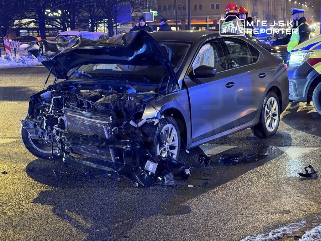 Czarny samochód osobowy z uszkodzoną przednią częścią stoi na ulicy, obok niego interweniują służby ratunkowe.
