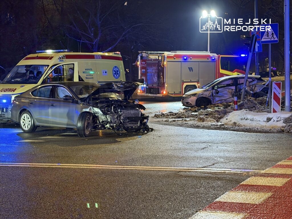 Dwa uszkodzone samochody na skrzyżowaniu, obok karetka pogotowia i wóz strażacki, nocą w miejskim otoczeniu.