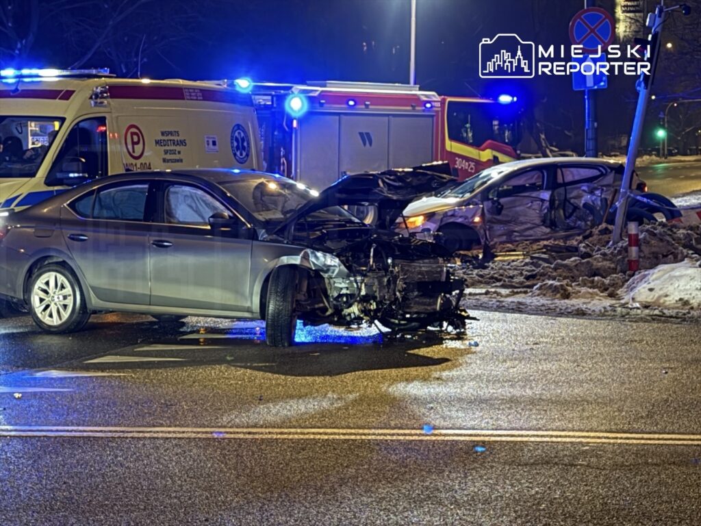 Zniszczony samochód osobowy na drodze, obok stoi karetka pogotowia oraz drugi uszkodzony pojazd.