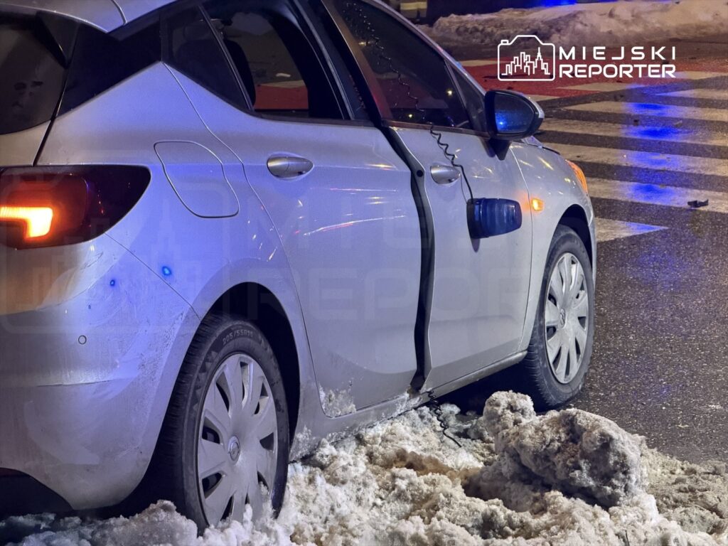 Uszkodzony samochód osobowy zaparkowany na ośnieżonej ulicy, z otwartymi drzwiami i widocznymi śladami kolizji.