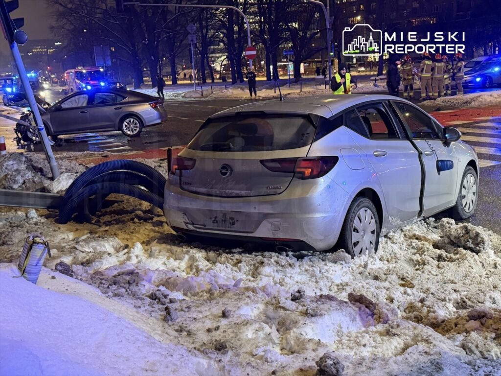 Samochód osobowy utknął w śniegu obok zniszczonego słupa, a w tle widać interwencję służb ratunkowych.
