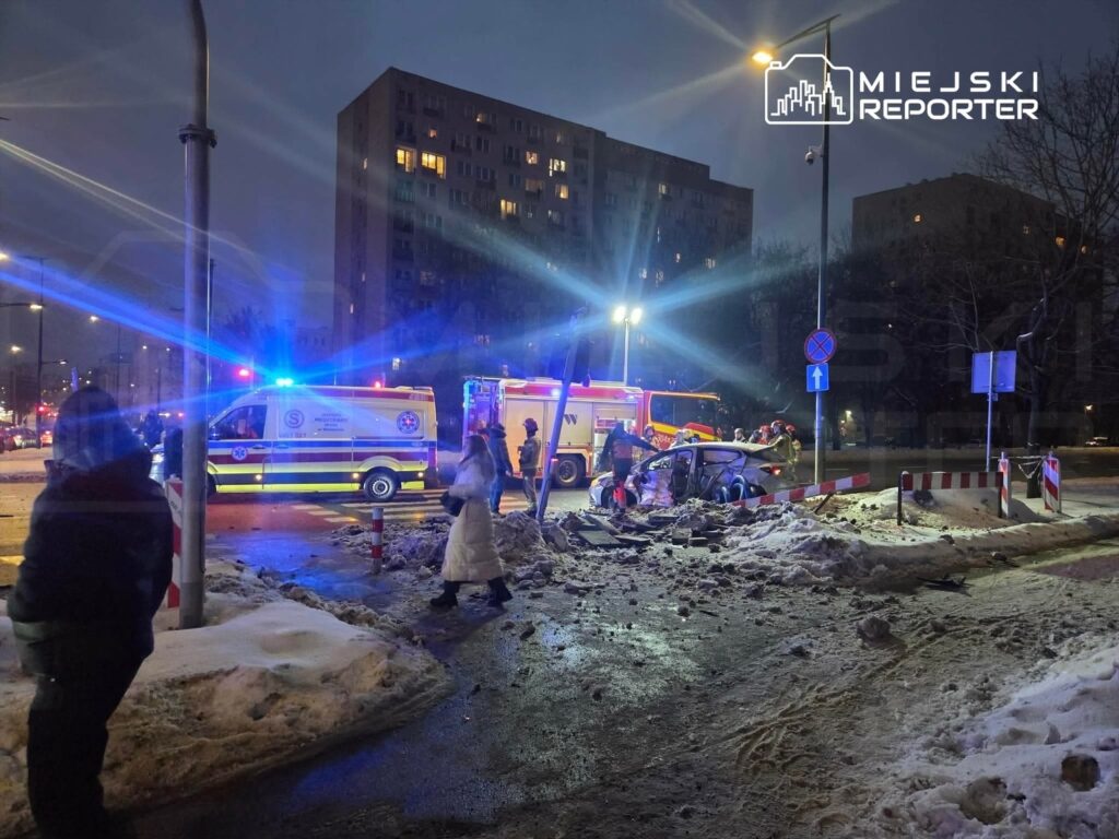 Na skrzyżowaniu w nocy, ratownicy medyczni udzielają pomocy przy rozbitym samochodzie, obok stoją karetki.