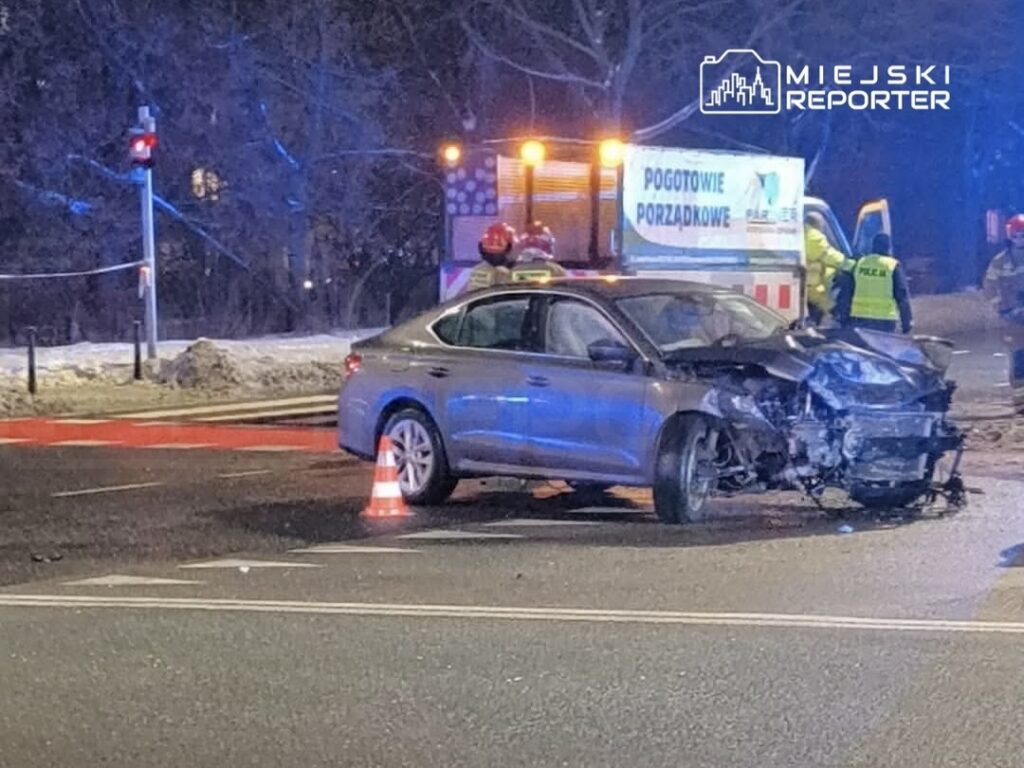 Uszkodzony samochód osobowy stoi na drodze, obok pracują służby ratunkowe oraz policja.