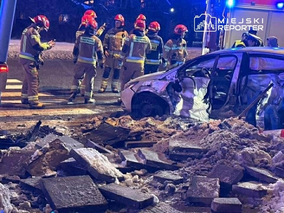 Grupa strażaków w hełmach omawia sytuację przy rozbitej osobówce na zaśnieżonej drodze, obok leżących płytek.