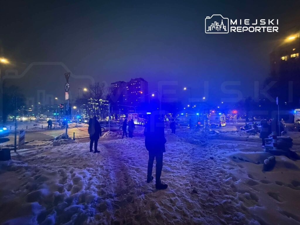 Grupa ludzi w ciemnych ubraniach stoi na ośnieżonej ulicy, otoczeni niebieskimi światłami radiowozów w nocy.