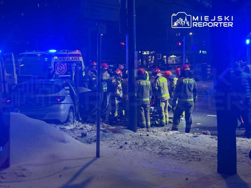 Grupa strażaków w hełmach i uniformach pracuje przy uszkodzonym samochodzie w oświetlonym niebieskimi światłami otoczeniu.