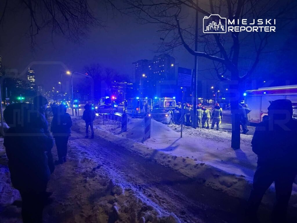 Grupa strażaków i ratowników medycznych pracuje w nocy na zaśnieżonej ulicy, otoczeni przez niebieskie światła pojazdów.