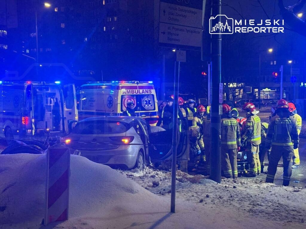 Grupa ratowników w hełmach i mundurach, udzielająca pomocy poszkodowanemu w wypadku samochodowym na ośnieżonej ulicy.