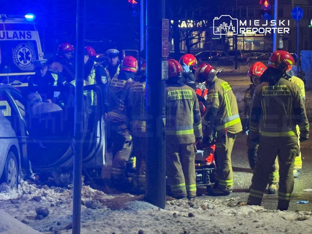 Grupa strażaków i ratowników medycznych udziela pomocy poszkodowanemu na ulicy w nocy, w otoczeniu pojazdów ratunkowych.