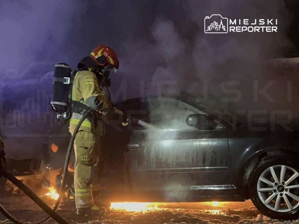 Strażak w kombinezonie ochronnym gasi płonący samochód, otoczony dymem i ogniem w nocy.