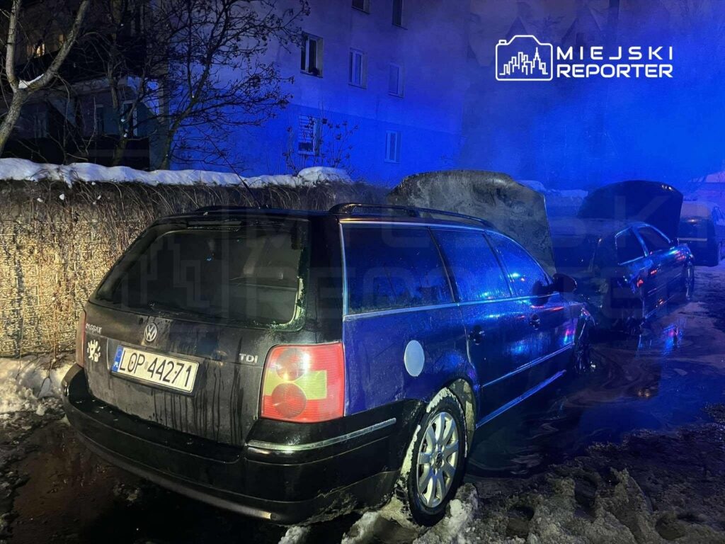 Czarny samochód osobowy zaparkowany na zaśnieżonej ulicy, obok inny pojazd z otwartą maską, w tle niebieskie światła.