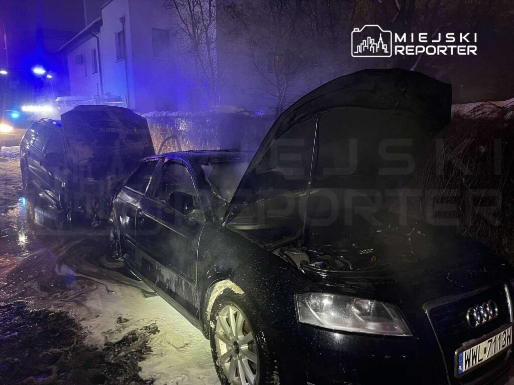 Dwa samochody osobowe, jeden z otwartą maską, stoją na ośnieżonej ulicy w nocy, otoczone dymem i światłami policyjnymi.