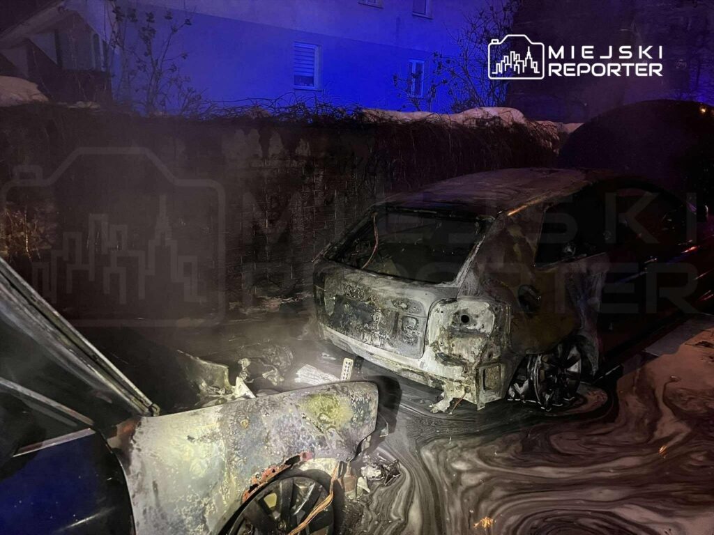 Spalone samochody stoją na zanieczyszczonym terenie, otoczone ciemnością i dymem.