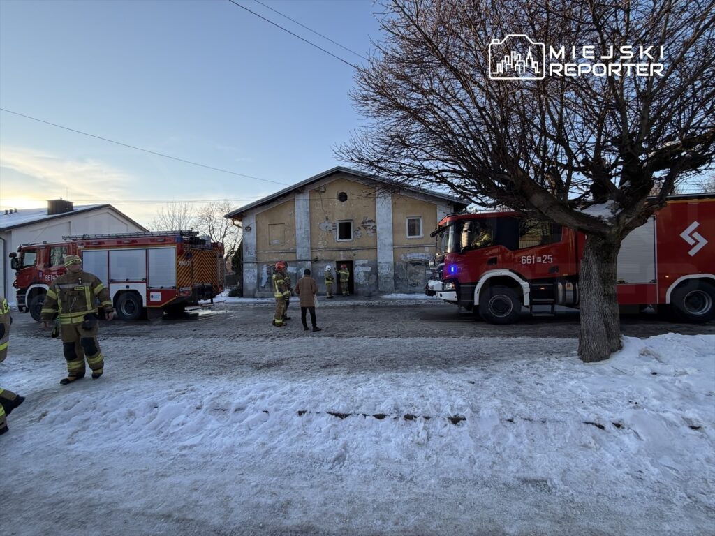 Grupa strażaków w mundurach rozmawia obok samochodów strażackich przed budynkiem w zimowej scenerii.