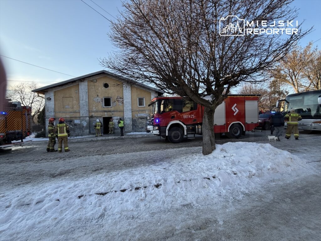 Grupa strażaków w hełmach pracuje przy budynku z uszkodzoną elewacją, obok stoją wozy strażackie na zaśnieżonej ulicy.