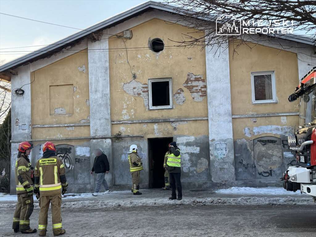 Grupa strażaków w mundurach rozmawia przed zniszczonym budynkiem, obok stoi mężczyzna w szarej bluzie.