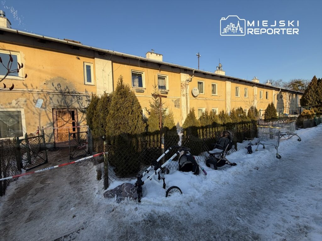 Zniszczony budynek mieszkalny z odrapaną elewacją, otoczony siatką, z wózkami dziecięcymi i zakupowymi na śniegu.