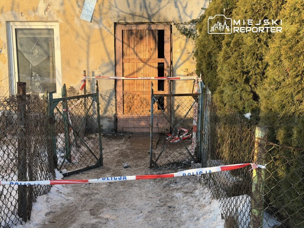 Zamknięta brama z siatki prowadząca do zniszczonego budynku, otoczona taśmą policyjną i zaroślami.