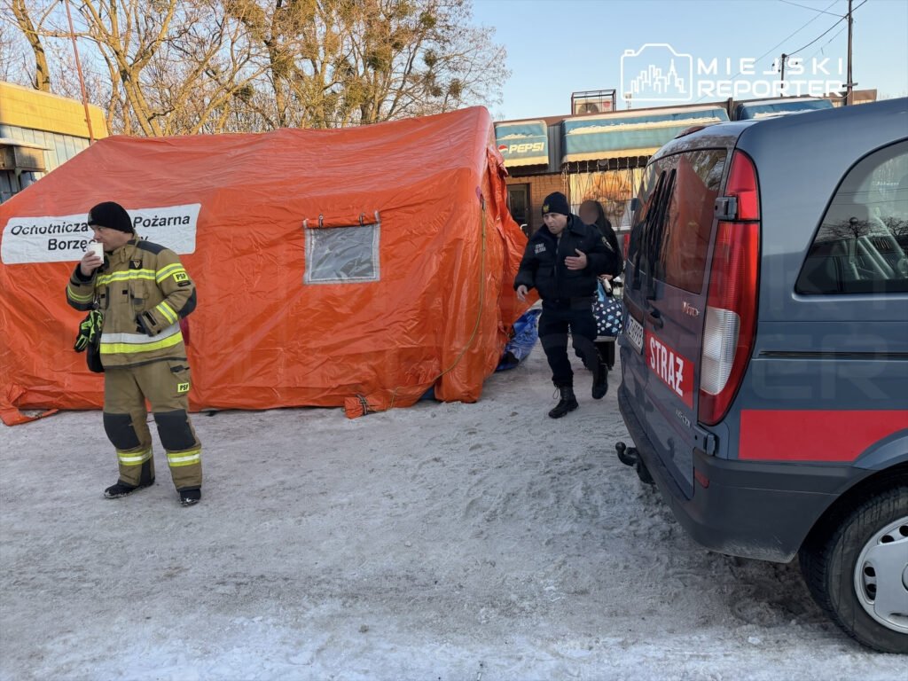 Strażak w mundurze stoi z kubkiem, obok pomarańczowego namiotu z napisem "Ochotnicza Straż Pożarna Borzęcin", w tle radiowóz strażacki.