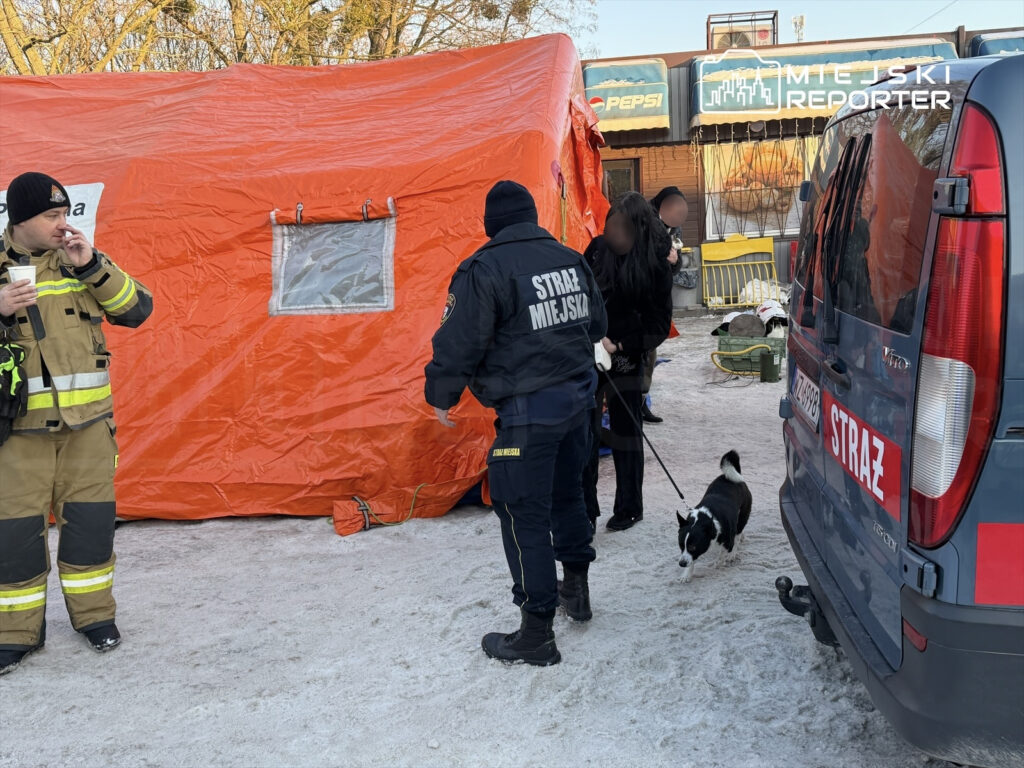 Strażacy i strażnicy miejscy pracują przy pomarańczowym namiocie, a jeden z nich prowadzi psa na smyczy.