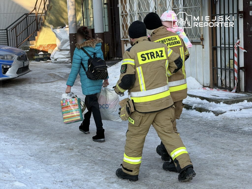 Dziecko w niebieskiej kurtce niesie torby, idąc obok strażaków w mundurach z odblaskowymi elementami w zimowym otoczeniu.