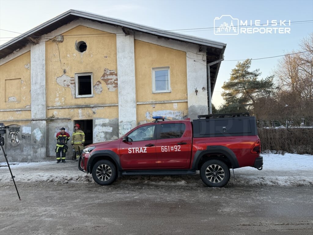 Strażacy w mundurach rozmawiają przed budynkiem z uszkodzoną elewacją, obok zaparkowany wóz strażacki.
