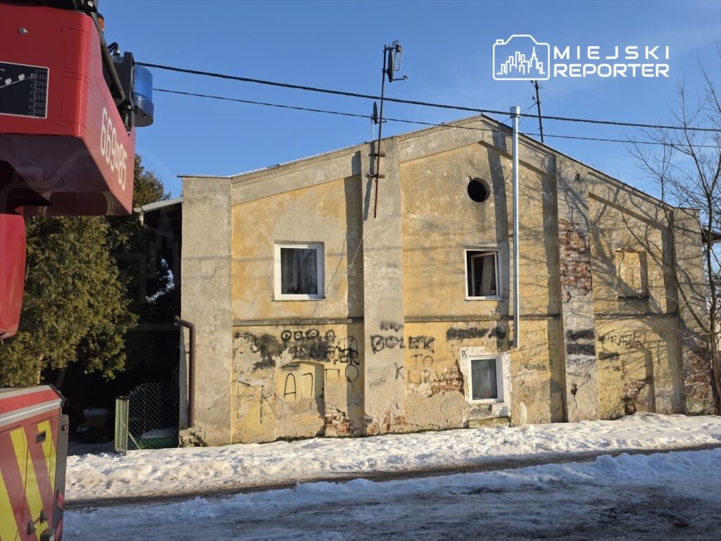 Czerwony wóz strażacki z sygnalizacją świetlną zaparkowany obok zaniedbanego budynku z graffiti na elewacji.