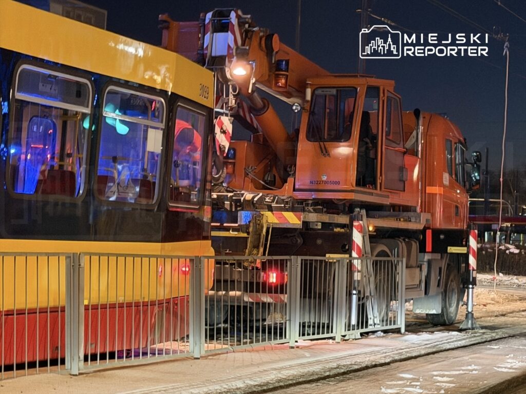 Tramwaj z żółtą i czarną malaturą zatrzymany obok pomarańczowego dźwigu, który podnosi elementy infrastruktury, w nocnej scenerii.