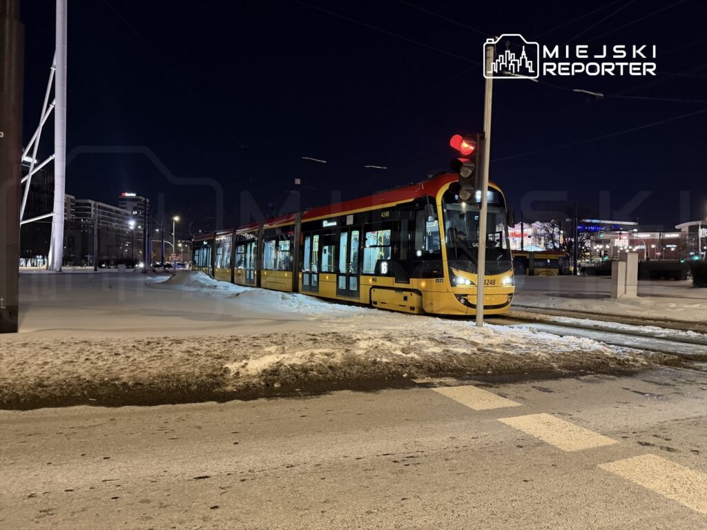 Tramwaj w nocy zatrzymany na przystanku, obok świecące lampy i ośnieżona ulica.
