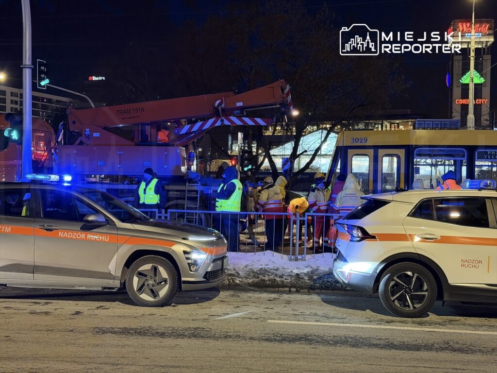 Grupa pracowników w odblaskowych kamizelkach naprawia tramwaj w nocy, obok stoją dwa radiowozy z włączonymi sygnałami świetlnymi.