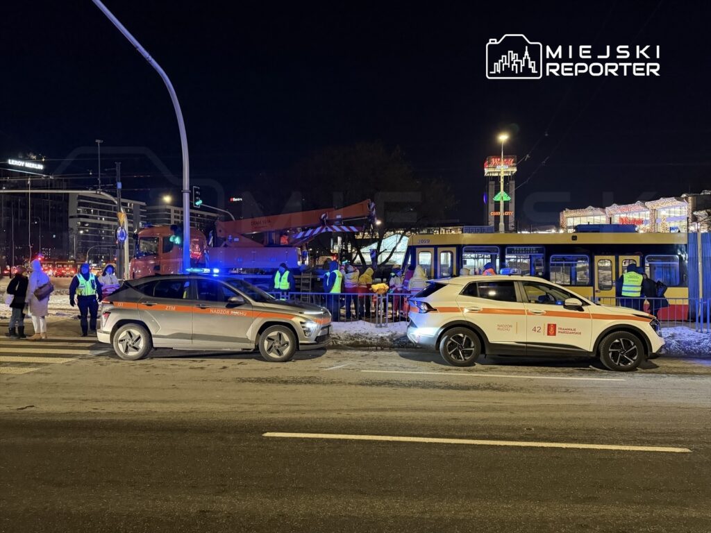 Grupa pracowników służb miejskich prowadzi prace przy tramwaju w nocnej scenerii, otoczona pojazdami służbowymi.
