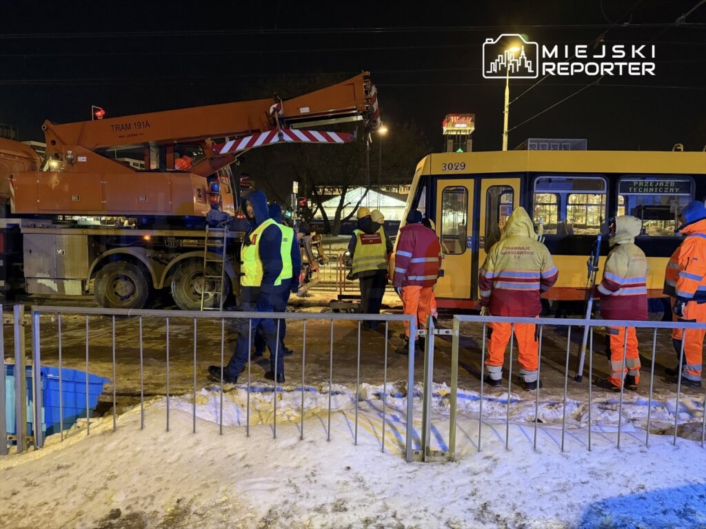 Pracownicy w odblaskowych kurtkach prowadzą prace przy tramwaju w nocy, obok podnośnika i ogrodzenia.