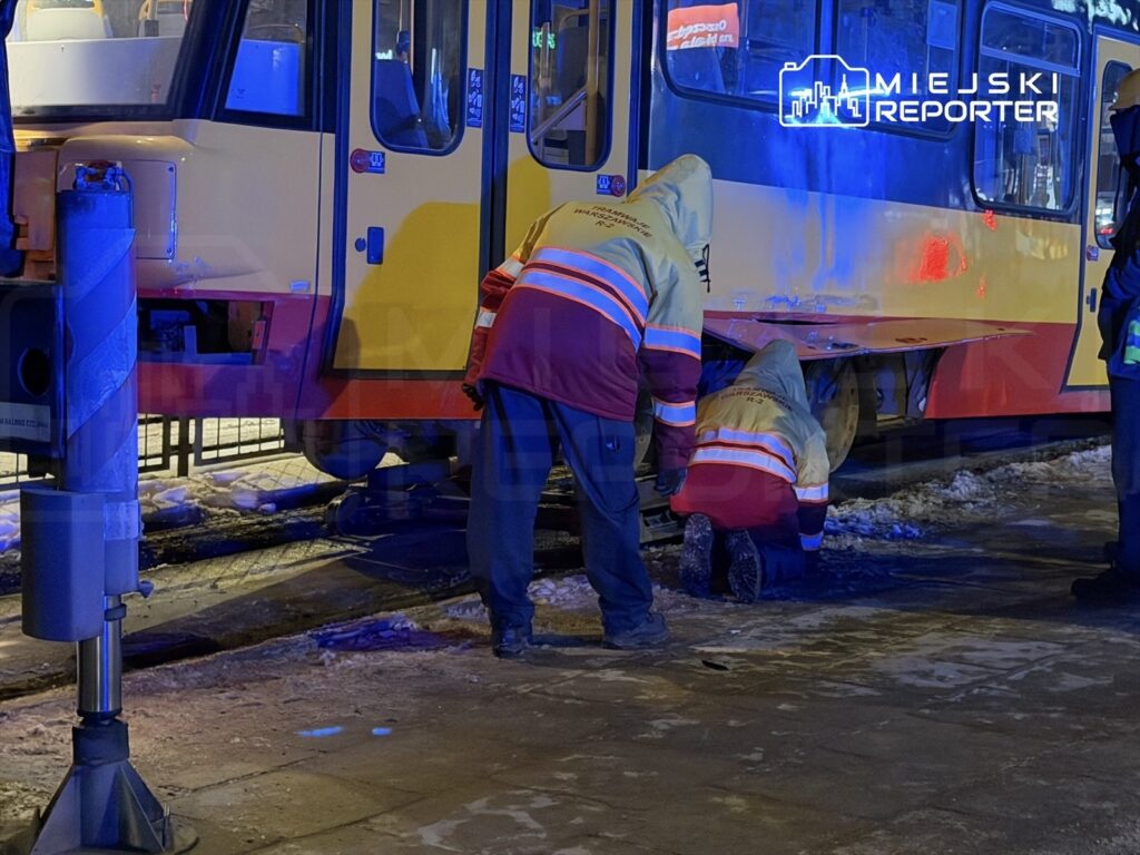 Pracownicy w odzieży roboczej naprawiają tramwaj na przystanku w zimowej scenerii, otoczeni śniegiem i sztucznym oświetleniem.